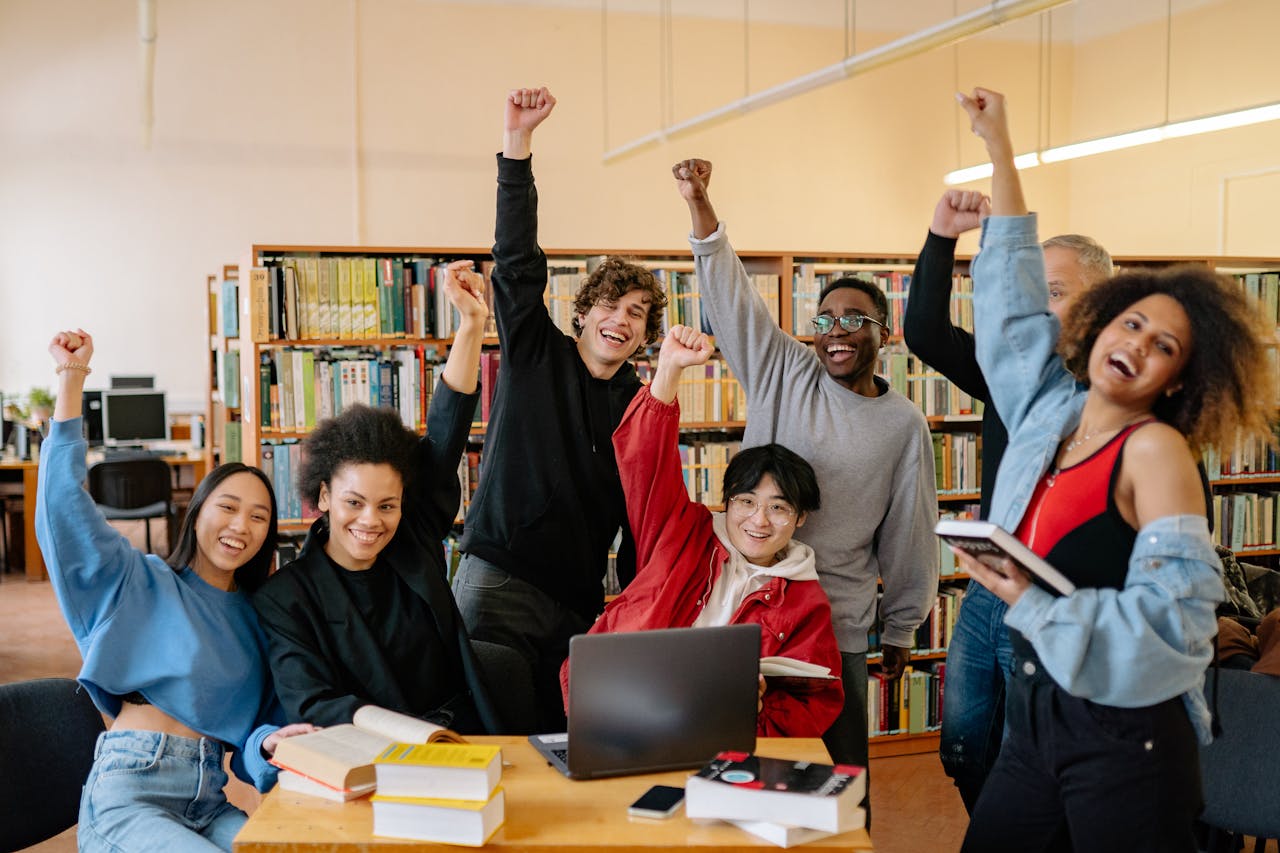 services-01 Happy diverse students in library raising hands, enjoying academic success.